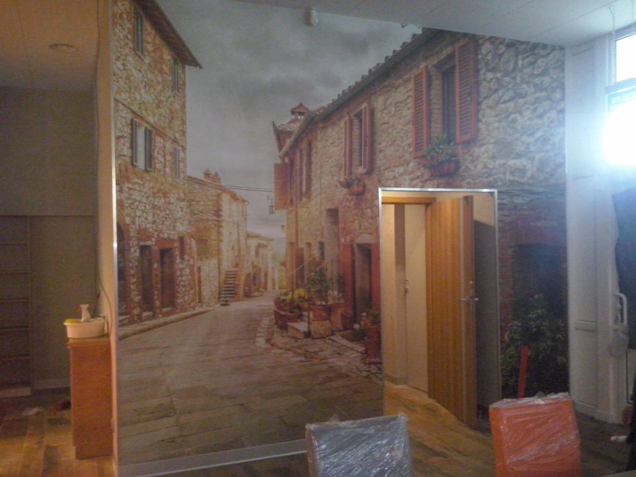 Fototapeta z motywem włoskiej uliczki na ścianie w pomieszczeniu. Widoczny fragment drzwi i okna. Częściowo zakryte meble i przedmioty w folii ochronnej.