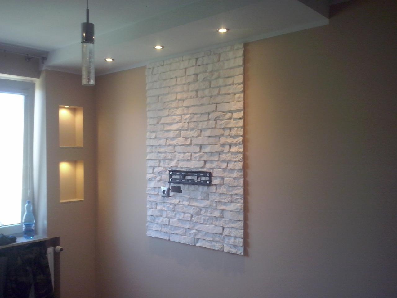 Beżowe wnętrze z dekoracyjną ścianą z białej cegły, na której zamontowano uchwyt na telewizor. Wnęki z oświetleniem i wisząca lampa dodają charakteru.