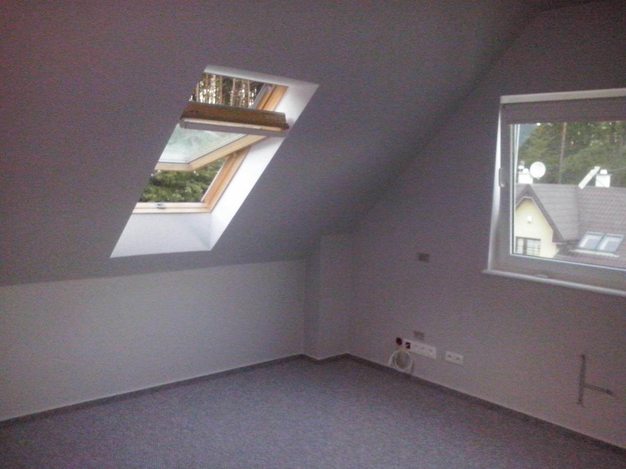 Poddasze z oknami dachowymi i standardowym, szare ściany po niedawnym malowaniu, widoczne gniazdka elektryczne i fragment grzejnika. Na podłodze położona wykładzina.