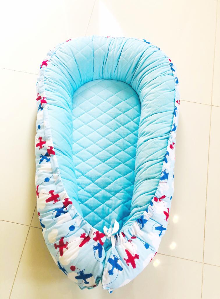Gotowe, pikowane gniazdko niemowlęce w błękitnym kolorze z motywem samolotów, leżące na jasnych płytkach podłogowych.