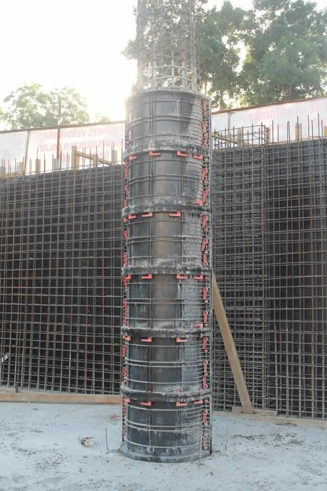 Szary, segmentowy szalunek kolumnowy z pomarańczowymi zamkami, otoczony zbrojeniem, na tle siatki zbrojeniowej i zieleni. Budowa w trakcie realizacji.
