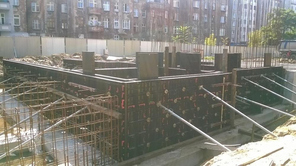 Zbrojenie i szalunek fundamentów w trakcie budowy, widoczne pręty zbrojeniowe, metalowe podpory i forma szalunkowa. W tle budynek mieszkalny i ogrodzenie.