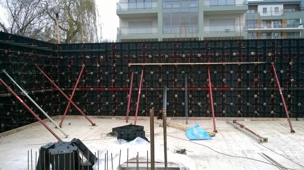 Szczegółowe ujęcie szalunków ściennych z podpórkami na budowie, widoczne elementy konstrukcyjne i zbrojenie, z budynkiem w tle.