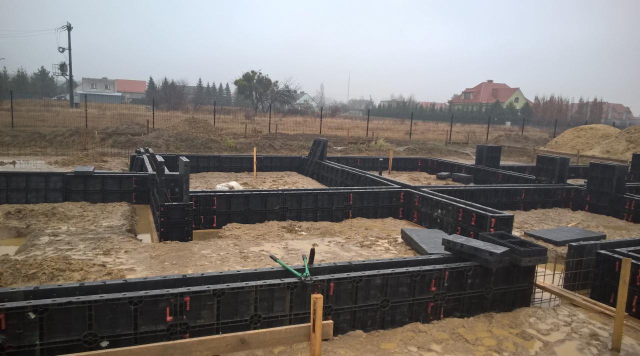 Szare, pochmurne niebo nad przygotowanym terenem pod fundamenty domu. Modułowe szalunki z tworzywa sztucznego ułożone w wykopie. W tle domy jednorodzinne.