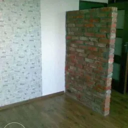 Wnętrze pokoju po remoncie: ściana z tapetą w drobny wzór, biała ściana i dekoracyjna ścianka z czerwonej cegły, parkiet. Widoczny fragment drzwi.