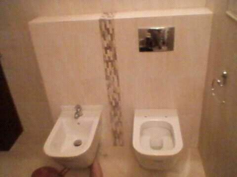 Wykończona łazienka w beżowych tonacjach: wisząca toaleta i bidet z baterią, oddzielone pionowym pasem mozaiki. Na ścianie chromowany przycisk spłuczki.