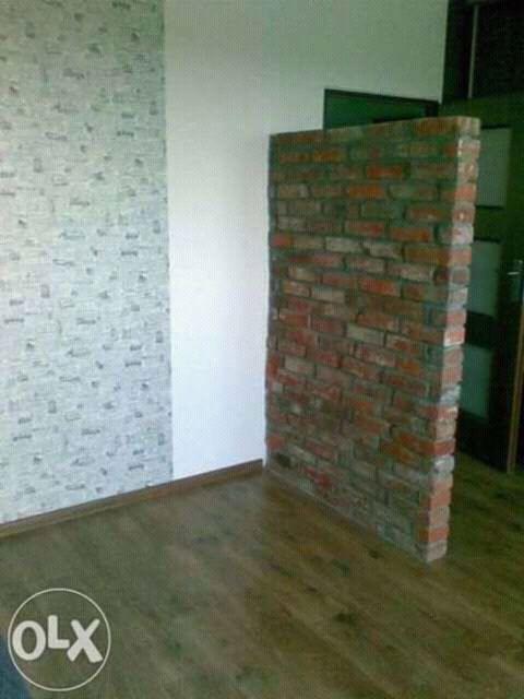 Wnętrze pokoju po remoncie: ściana z tapetą w drobny wzór, biała ściana i dekoracyjna ścianka z czerwonej cegły, parkiet. Widoczny fragment drzwi.