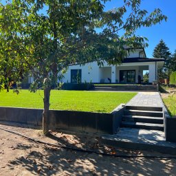 M.R. Drobet - Nowoczesny dom z ogrodem, schody i ścieżka wyłożone szarą kostką brukową, widoczny fragment prac ziemnych z kopiącym mężczyzną w tle.