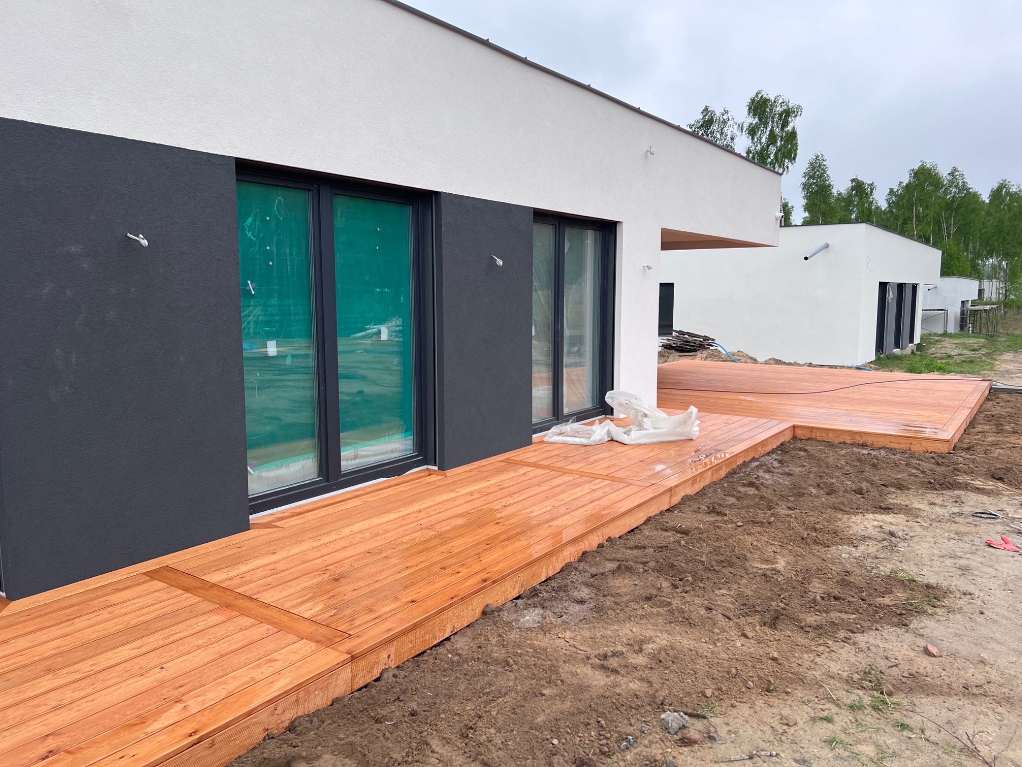 Nowoczesny dom z drewnianym tarasem w trakcie budowy, widoczne zabezpieczone okna i przygotowany teren wokół.