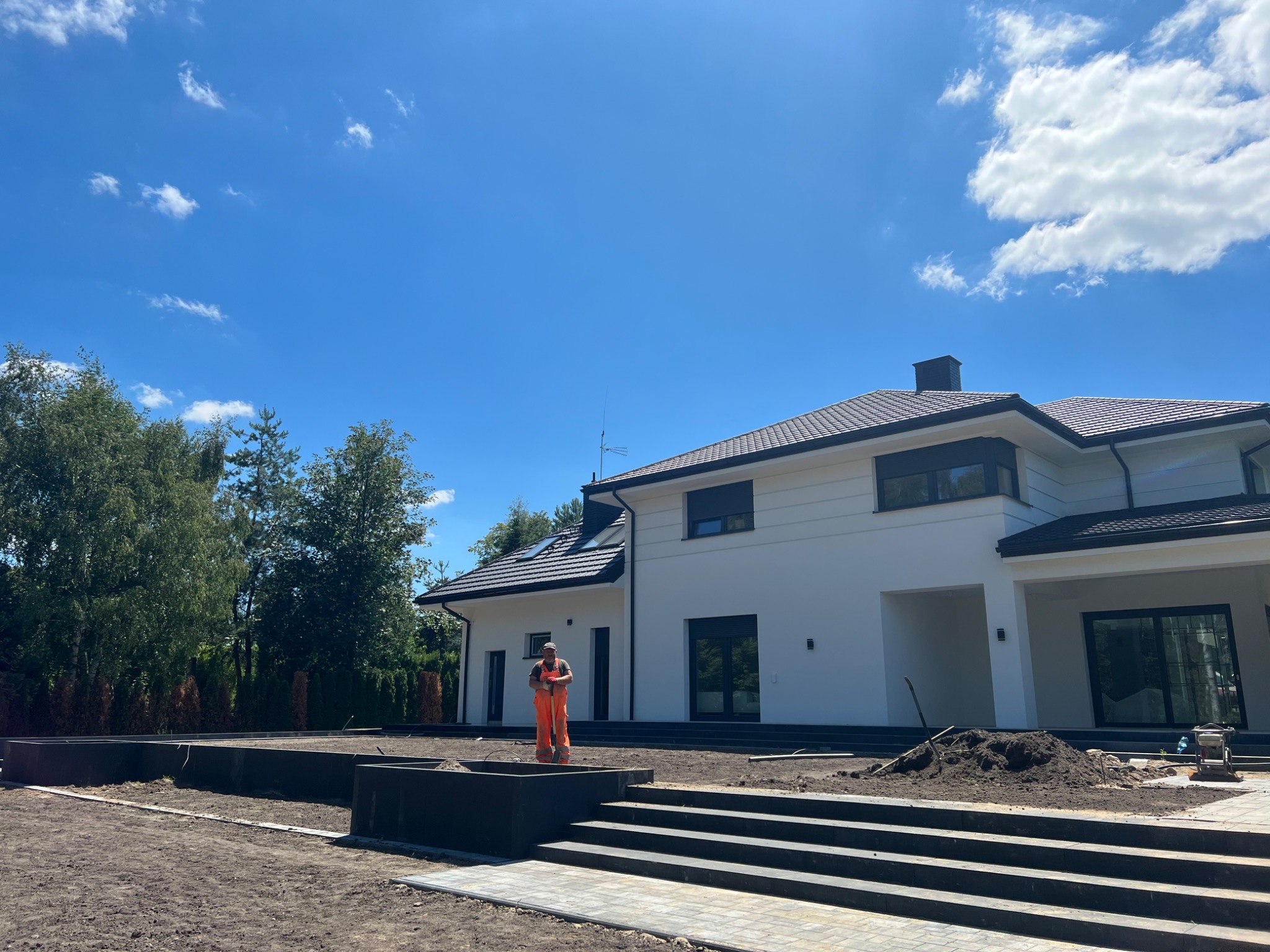 Pracownik w pomarańczowym kombinezonie z łopatą na tle nowoczesnego, białego domu w trakcie prac ziemnych, widoczne schody z kostki brukowej i przygotowane rabaty.