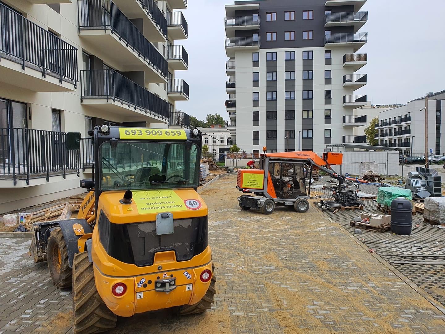 Żółta mini ładowarka z napisem 'brukartstwo niwelacja terenu' na tle nowo wybudowanych apartamentowców i częściowo ułożonej kostki brukowej z piaskiem, obok pomarańczowa maszyna do układania kostki.