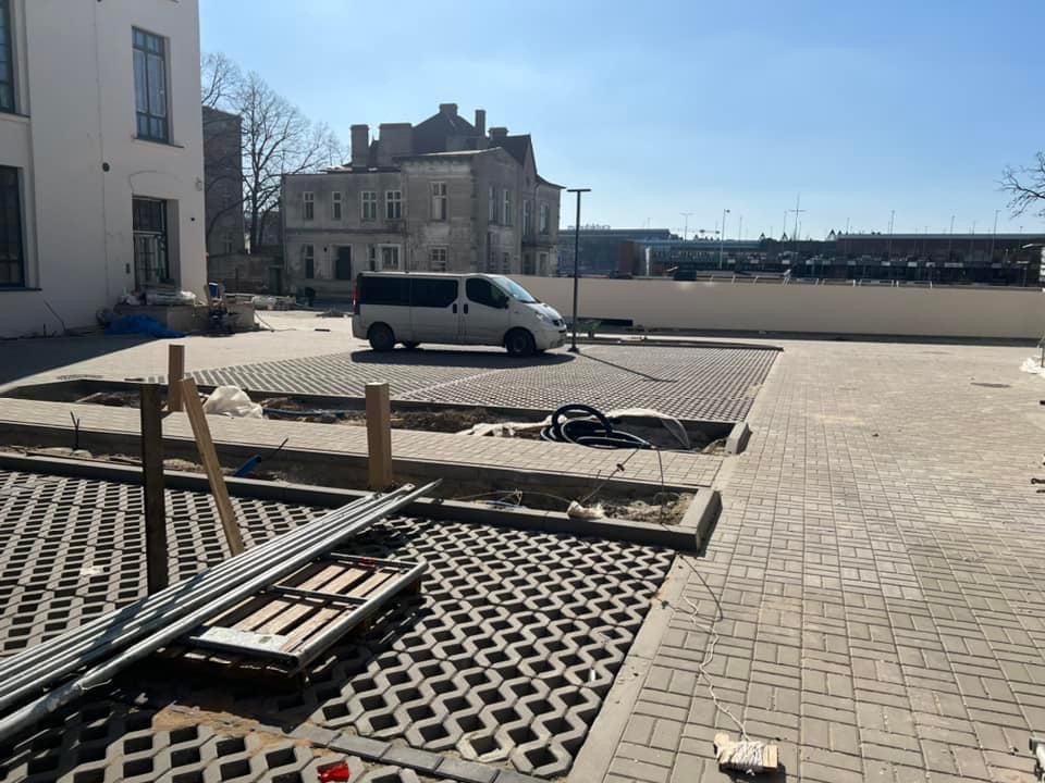 Fragment placu z ażurowych płyt betonowych w trakcie realizacji, widoczne przygotowane podłoże, krawężniki, rury i zaparkowany van w tle.