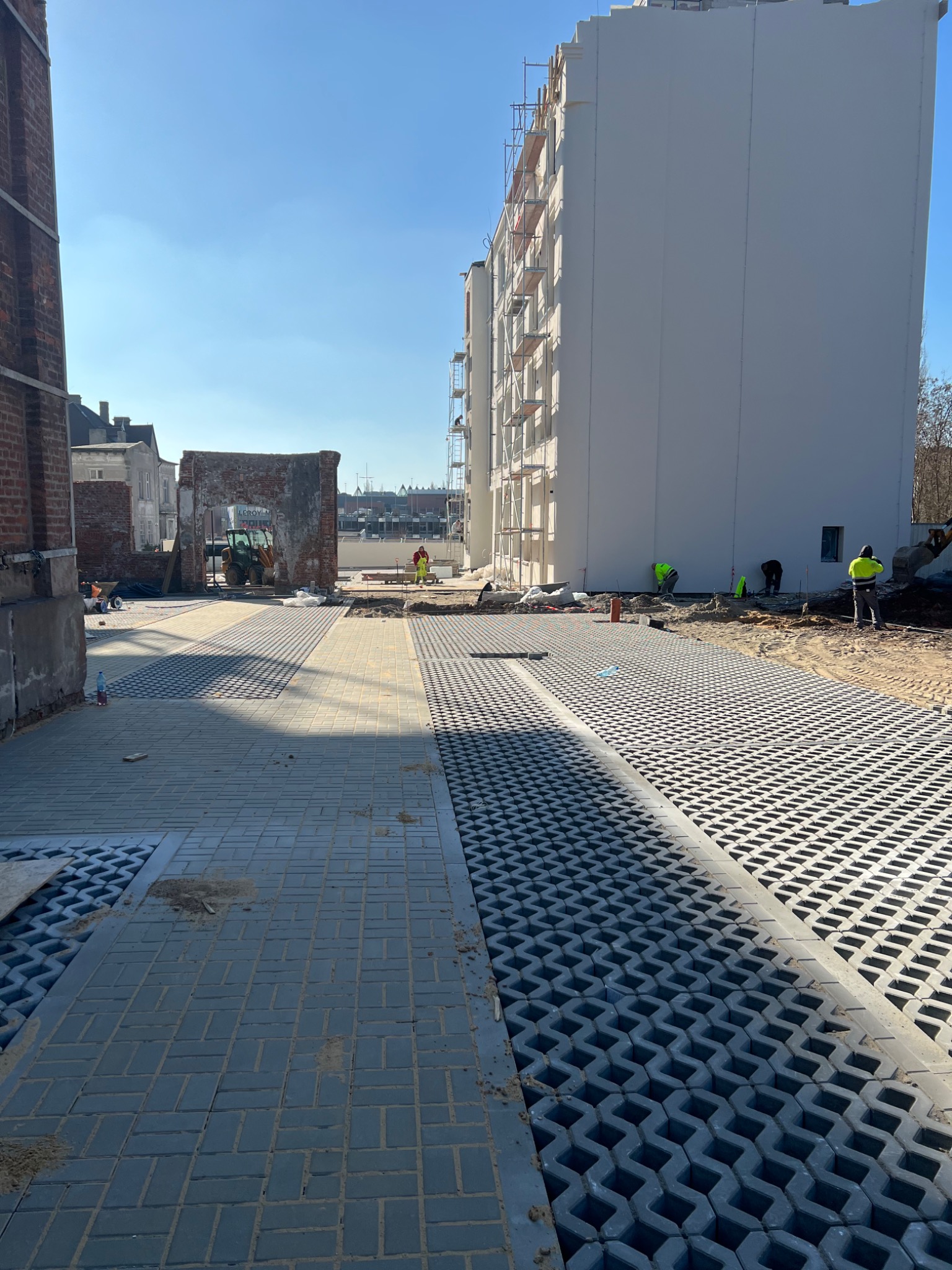 Obszar budowy z częściowo ułożoną nawierzchnią z kostki brukowej i ażurowych płyt betonowych, widoczni pracownicy w odblaskowych ubraniach oraz maszyny budowlane w tle.