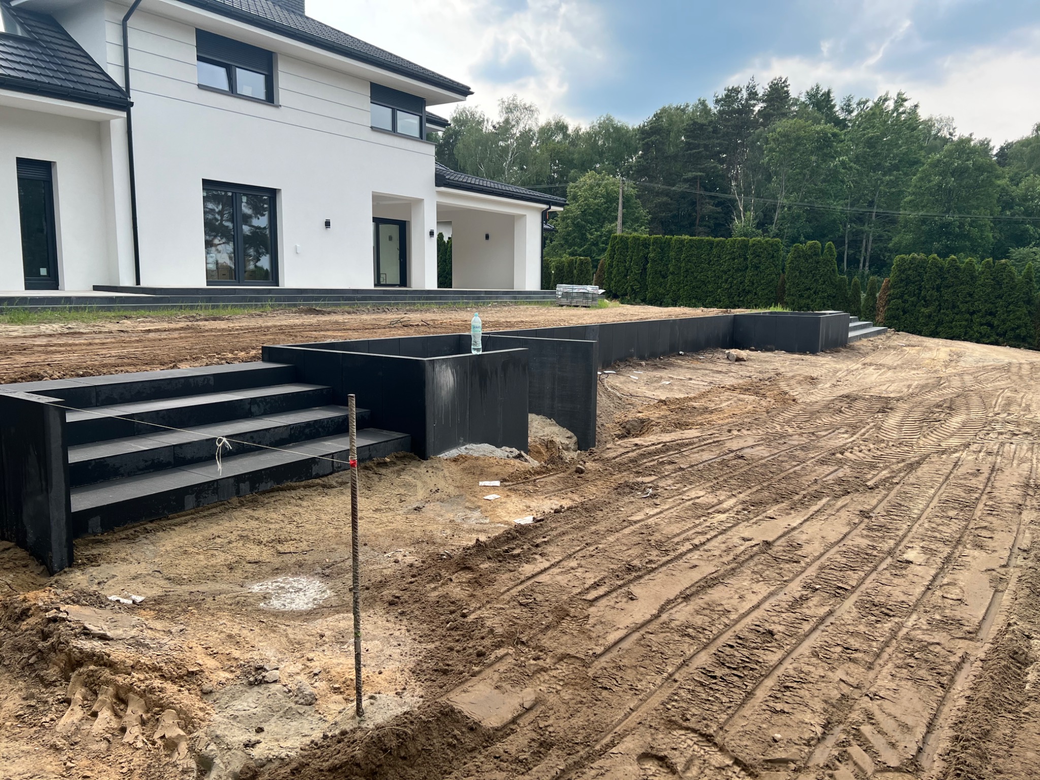 Nowoczesny dom z czarnymi schodami i murkami z kostki, teren w trakcie przygotowania, widoczne ślady opon i butelka wody mineralnej.