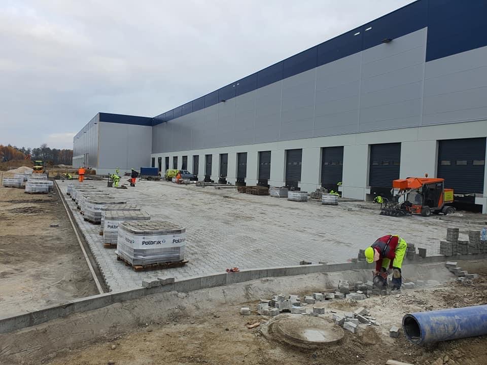 Prace brukarskie przed budynkiem magazynowym: robotnicy w odblaskowych ubraniach układają szarą kostkę, palety z kostką Polbruk ustawione w rzędzie, w tle maszyna do zagęszczania gruntu.