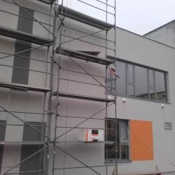 SPEC-THERM Grzegorz Piwowarski - Budynek w trakcie prac elewacyjnych, widoczne rusztowania, częściowo otynkowana ściana z pomalowanym na pomarańczowo fragmentem wokół okna, oraz paczka materiałów izolacyjnych na rusztowaniu.