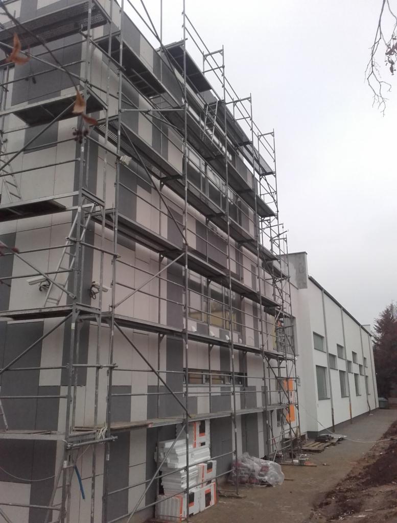 Budynek w trakcie ocieplania elewacji, widoczne rusztowania, częściowo ocieplona ściana z szarymi i białymi panelami, stosy materiałów izolacyjnych u podstawy rusztowania.