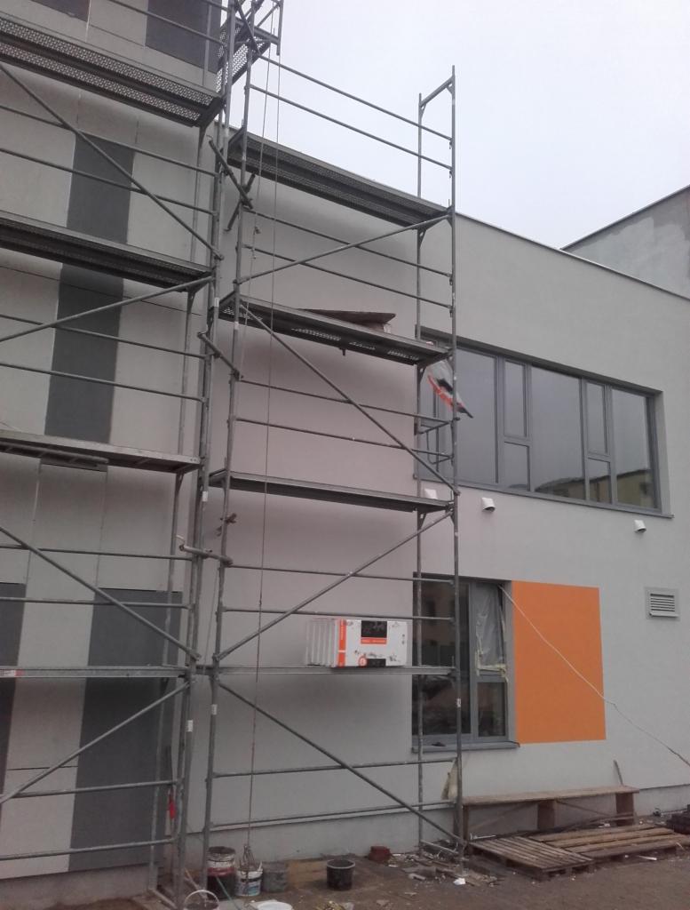 Budynek w trakcie prac elewacyjnych, widoczne rusztowania, częściowo otynkowana ściana z pomalowanym na pomarańczowo fragmentem wokół okna, oraz paczka materiałów izolacyjnych na rusztowaniu.