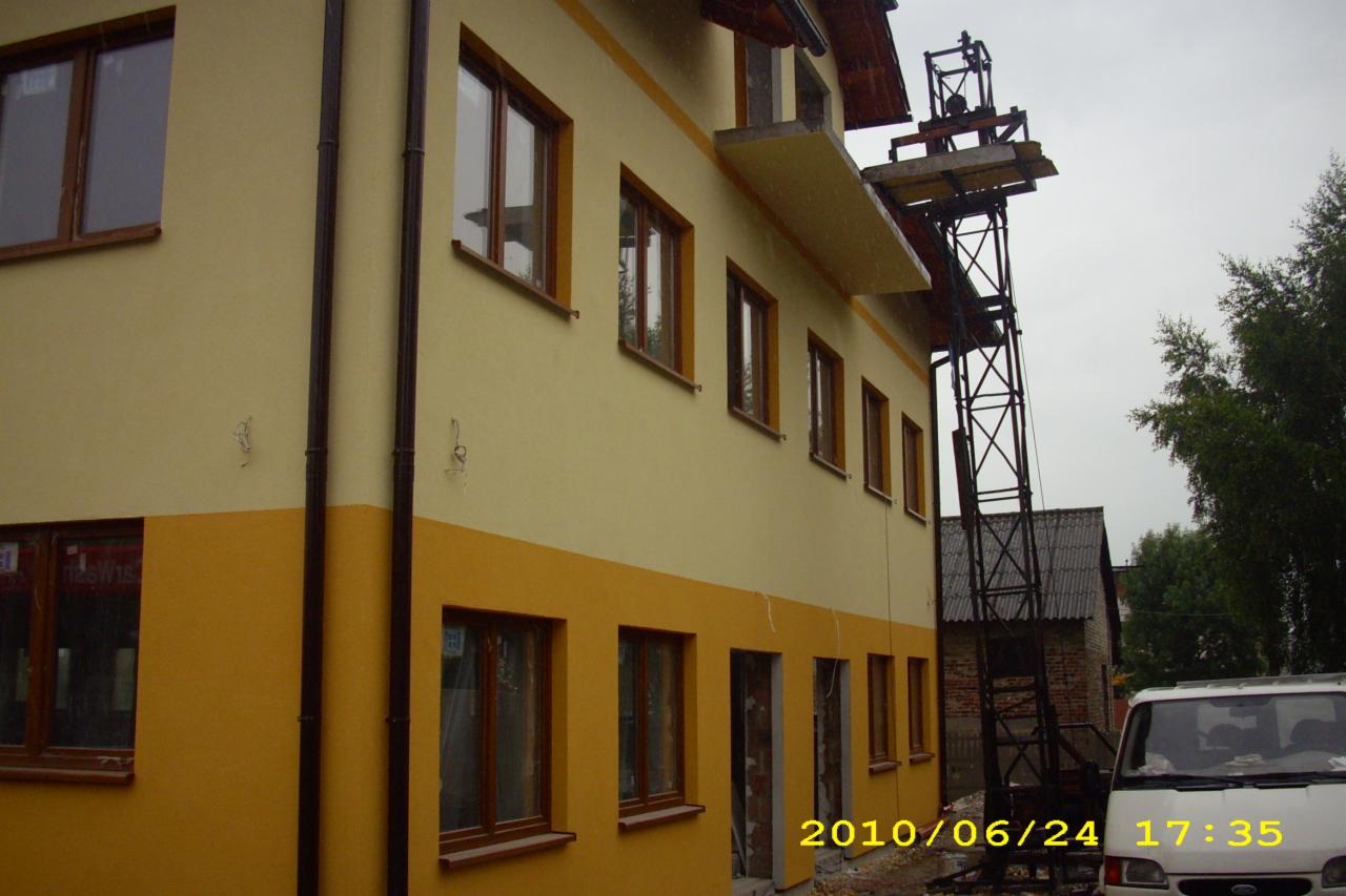 Dwukolorowa elewacja budynku w trakcie prac, z widoczną windą budowlaną i niezabezpieczonymi otworami okiennymi, wykonana w odcieniach żółci i pomarańczy, z datą wykonania zdjęcia w prawym dolnym...