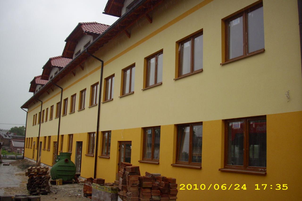 Dwukolorowa elewacja budynku po remoncie, z widocznymi stertami cegieł i zielonym zbiornikiem na trawniku, uchwycona w pochmurny dzień.