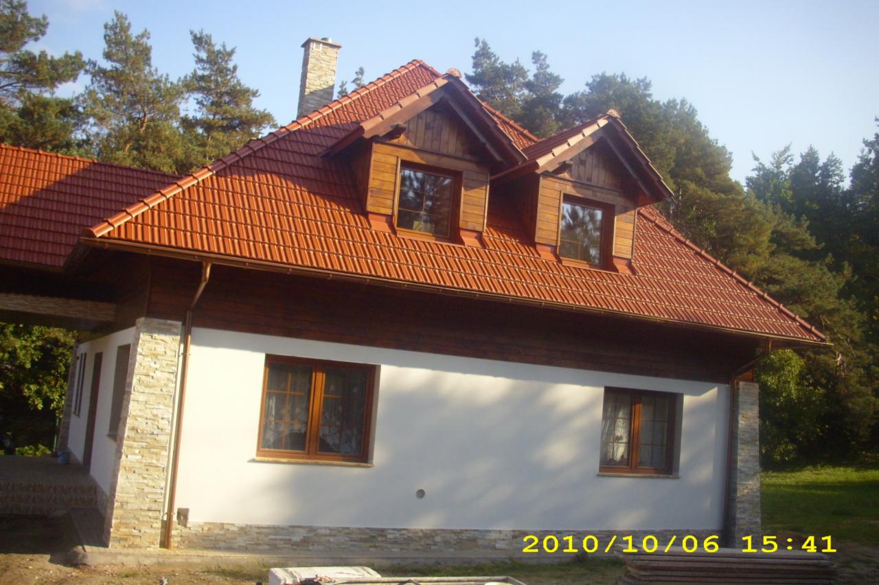 Dom jednorodzinny z czerwoną dachówką i drewnianymi oknami, wykończony kamieniem dekoracyjnym u podstawy, otoczony drzewami w słoneczny dzień.