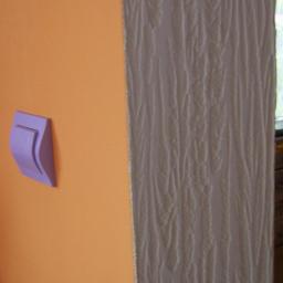 Loukas - Fragment pomarańczowej ściany z fioletowym włącznikiem światła, sąsiadującej z pionowym pasem ściany pokrytej tapetą o fakturze przypominającej korę drzewa.