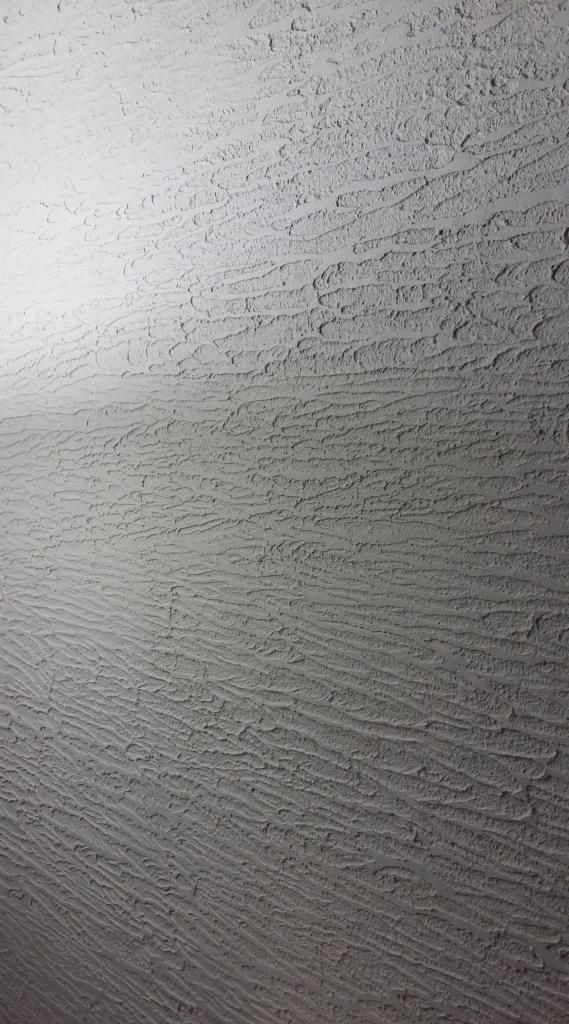 Szara ściana z dekoracyjnym, strukturalnym tynkiem w stylu baranka, oświetlona od góry, z widoczną fakturą i cieniem.