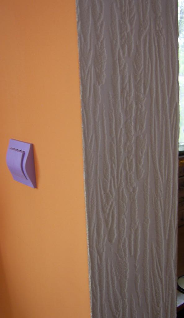 Fragment pomarańczowej ściany z fioletowym włącznikiem światła, sąsiadującej z pionowym pasem ściany pokrytej tapetą o fakturze przypominającej korę drzewa.