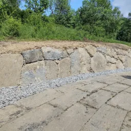 Kamienny murek oporowy, zabezpieczający skarpę porośniętą zielenią, wykończony grysem, z wybrukowanym chodnikiem z betonowych płyt.