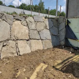 Kamienny mur oporowy w trakcie budowy, widoczna izolacja fundamentów i błotniste podłoże