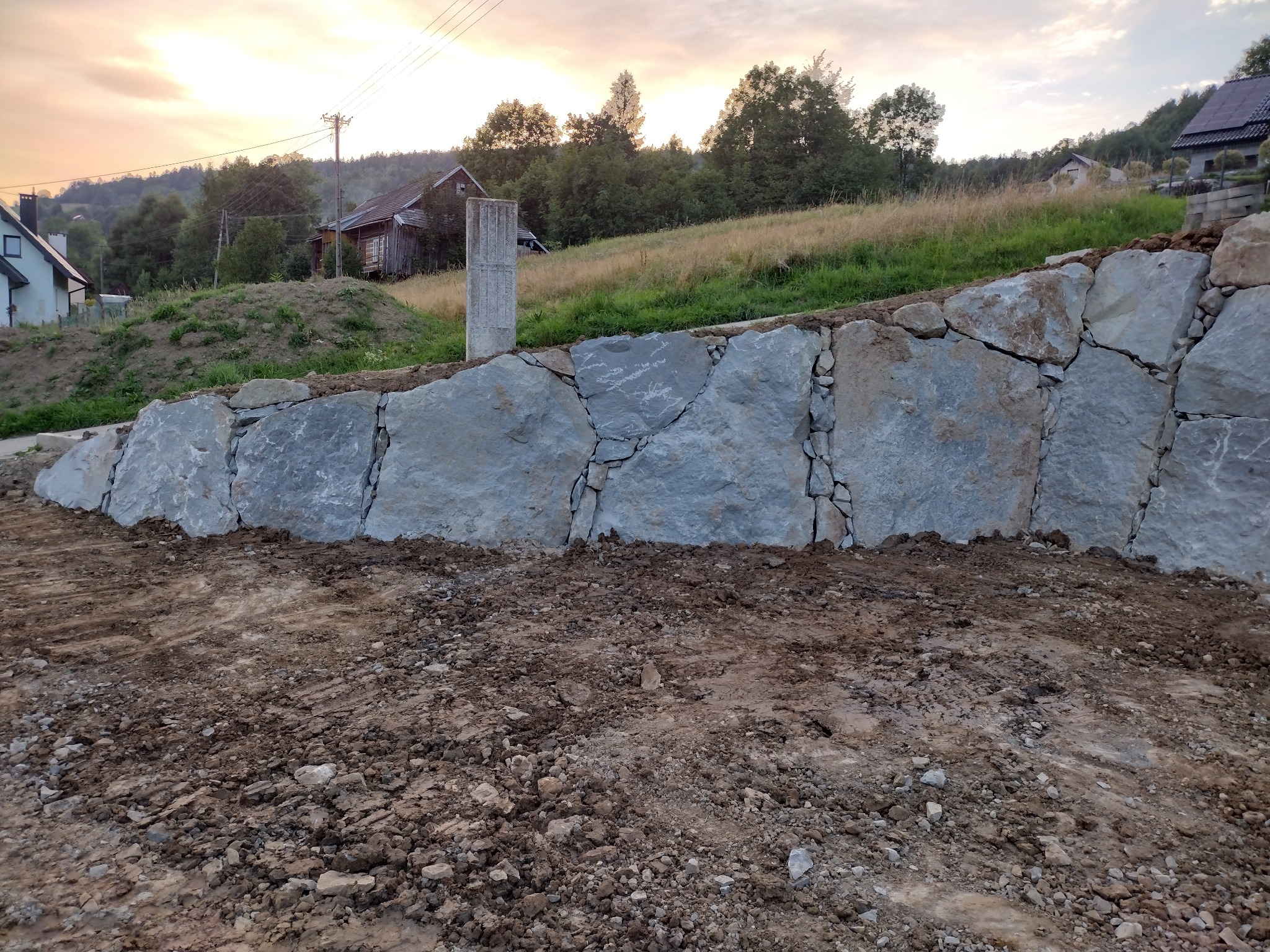 Kamienny mur oporowy na nierównym terenie, z widocznymi szczelinami i ziemią u podnóża, w tle wiejski krajobraz o zmierzchu.