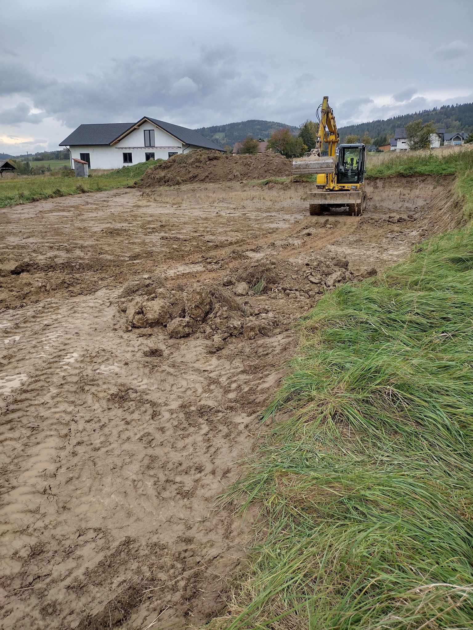 Żółta koparka w trakcie prac ziemnych na placu budowy, widoczne świeże ślady opon w błotnistym terenie, w tle dom jednorodzinny i zalesione wzgórza pod pochmurnym niebem.