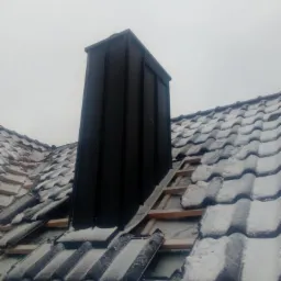 Czarny komin z blachy na dachu pokrytym częściowo śniegiem i dachówką, widoczne fragmenty konstrukcji drewnianej.