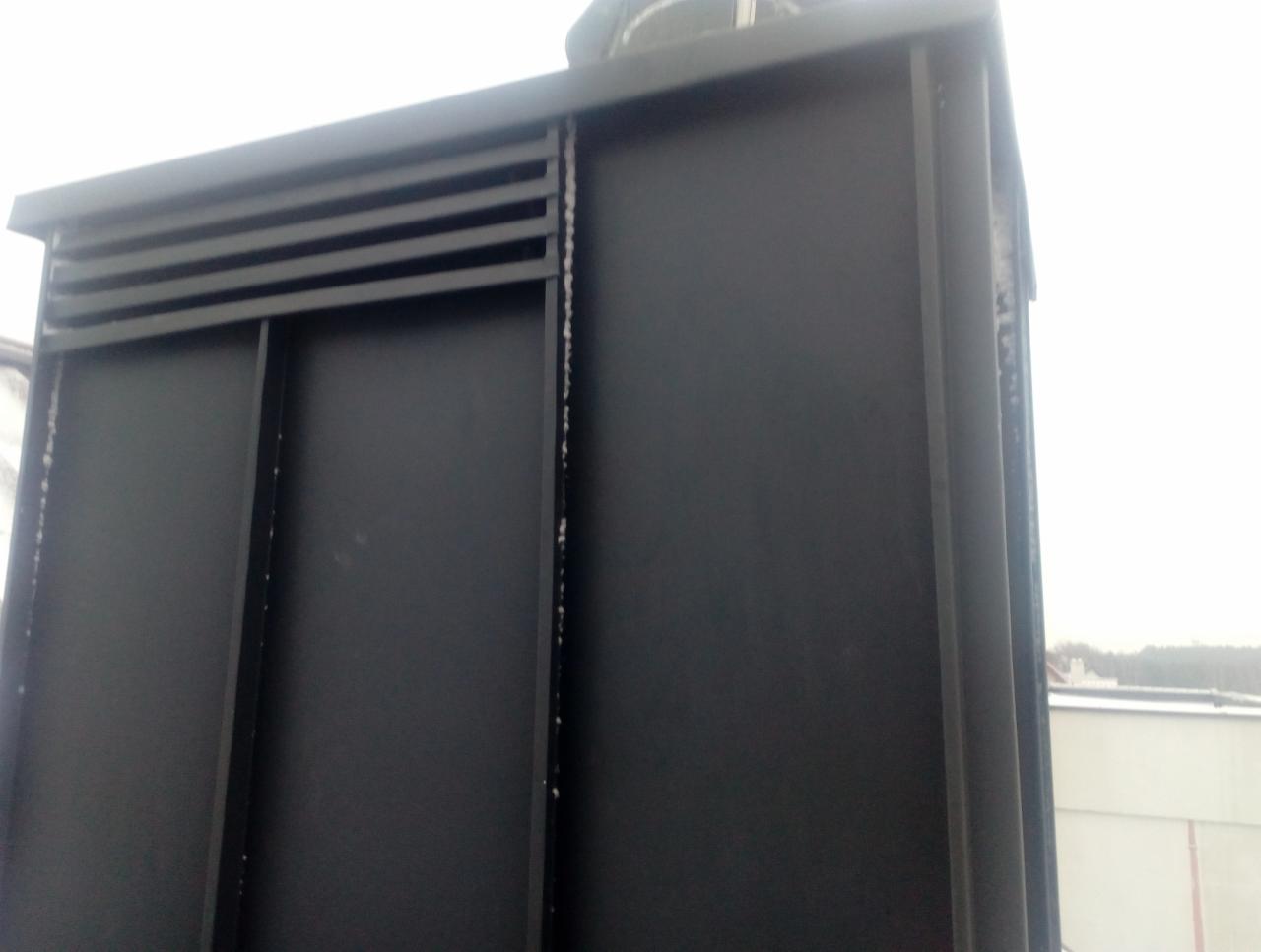 Fragment czarnej obudowy komina na dachu, z widocznymi poziomymi żaluzjami wentylacyjnymi i śladami zabrudzeń po prawej stronie, na tle pochmurnego nieba.