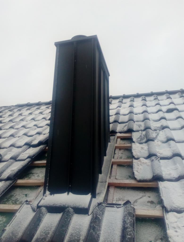 Czarny, metalowy komin na dachu pokrytym dachówką, częściowo ośnieżonym, widoczne elementy konstrukcji dachu w trakcie montażu.