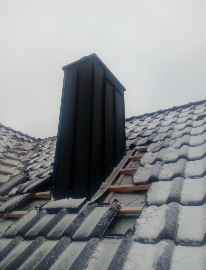 Czarny komin z blachy na dachu pokrytym częściowo śniegiem i dachówką, widoczne fragmenty konstrukcji drewnianej.