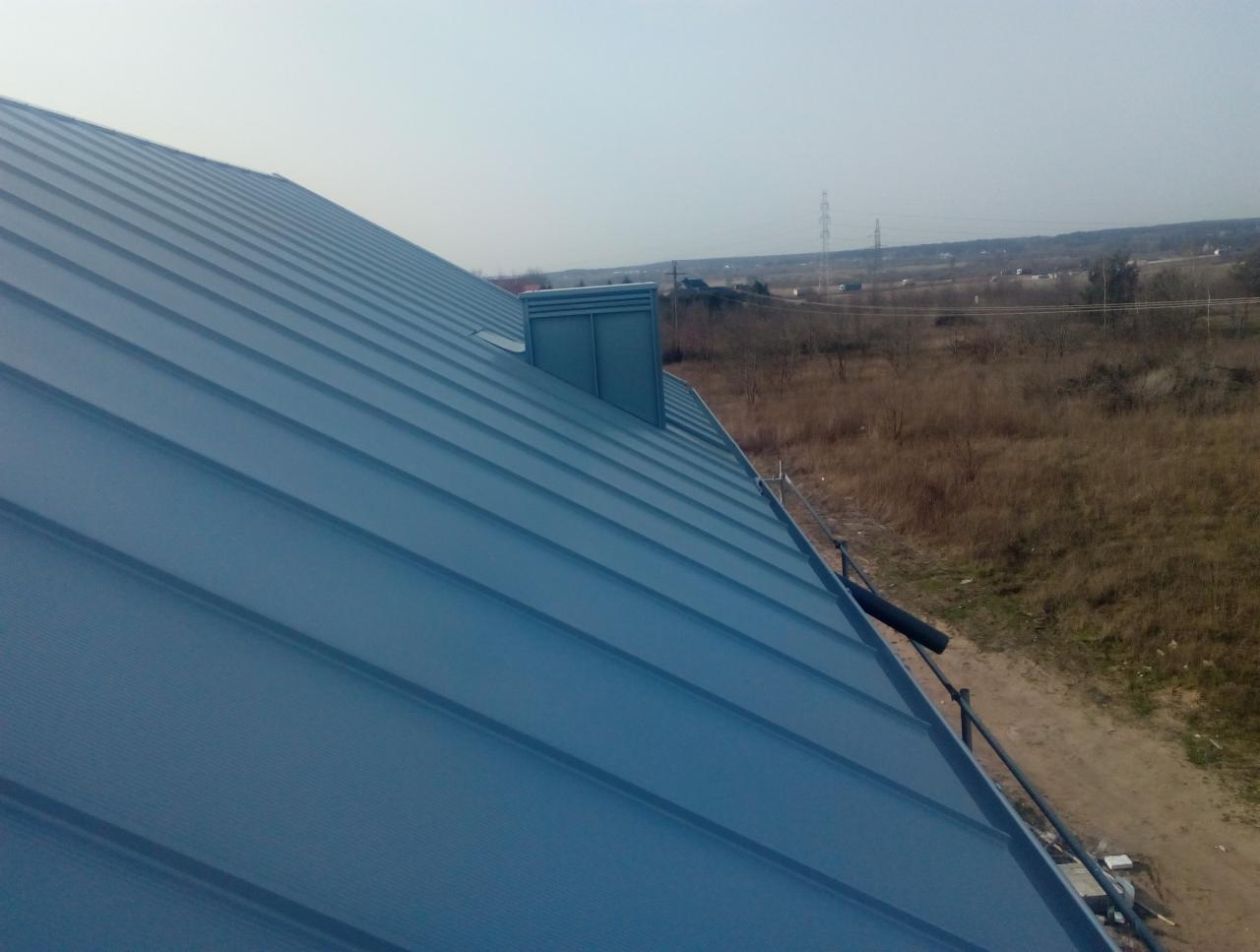 Niebieski dach z blachy trapezowej z kominkiem wentylacyjnym i rynną, widok z góry na tle jesiennego krajobrazu.