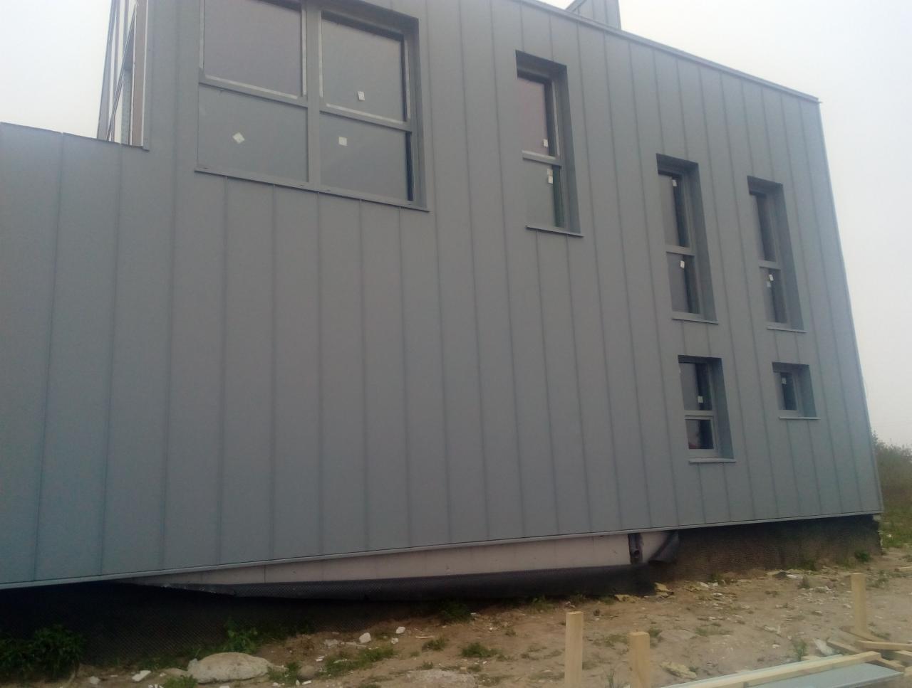 Nowoczesny budynek mieszkalny z szarą blachą na elewacji i dachu, widok z dołu, widoczne okna z szarą ramą i elementy konstrukcyjne pod elewacją.