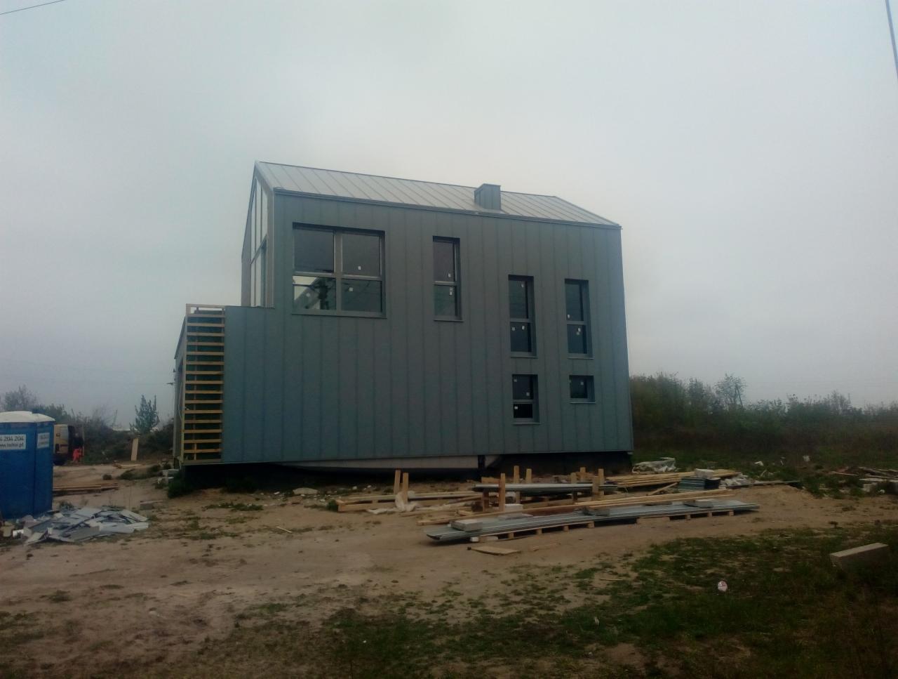 Nowoczesny dom w trakcie budowy z szarym metalowym poszyciem i dachem, widoczne rusztowania i materiały budowlane na placu budowy w mglisty dzień.