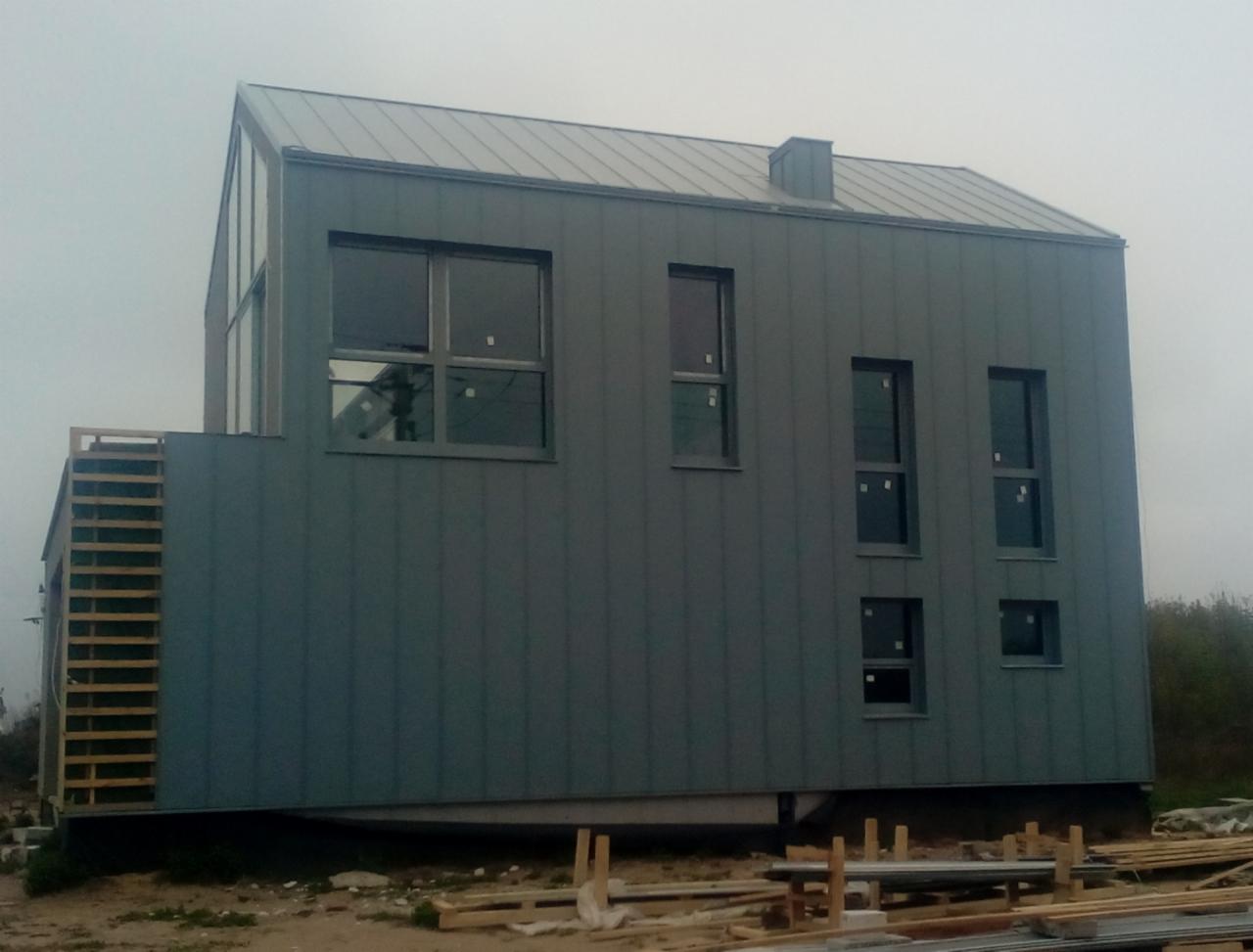 Nowoczesny dom z szarej blachy trapezowej, widoczne okna z szarymi ramami, fragment drewnianej konstrukcji balkonu i elementy składowane na ziemi.