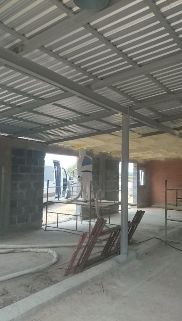 Ocieplanie pianką PUR stropu w surowym budynku gospodarczym, pracownik w kombinezonie ochronnym aplikuje pianę, widoczny agregat i przewody, surowy beton i cegły.