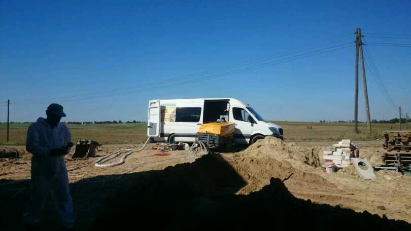 Pracownik w kombinezonie ochronnym kontroluje aplikację pianki PUR z zaparkowanego białego furgonetki na placu budowy, widoczny agregat i przewody.