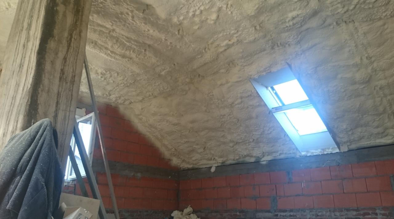 Poddasze z oknem dachowym po nałożeniu izolacji natryskowej, widoczne ceglane ściany i betonowy filar z zaciekami.