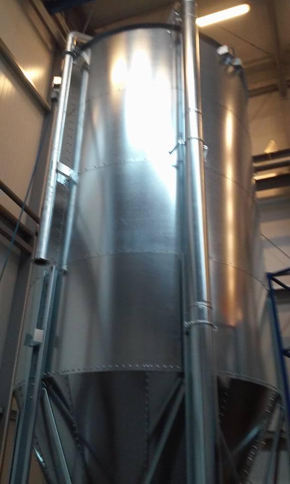 Błyszczący, metalowy silos z rurami i drabinką w hali produkcyjnej. Widok z dołu podkreśla jego wysokość i konstrukcję. Oświetlenie eksponuje metaliczną powierzchnię.