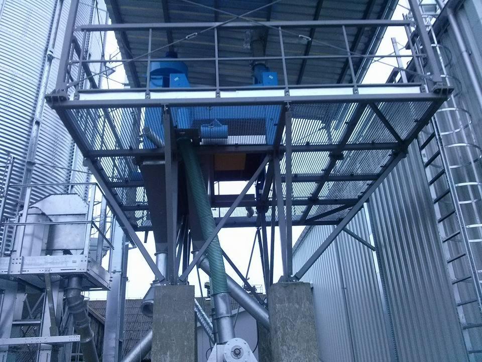 Stalowa konstrukcja wsporcza z platformą kratową, element instalacji przemysłowej z silosami w tle, widok z dołu.