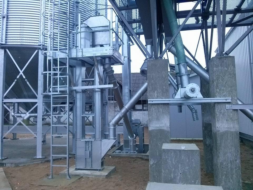 Srebrna konstrukcja stalowa z drabinką i rurami na zewnątrz, prawdopodobnie część większej instalacji przemysłowej lub rolniczej.