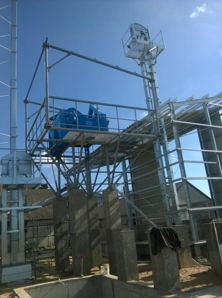 Wznoszenie stalowej konstrukcji hali z niebieskim elementem maszynerii na platformie, widok z dołu na tle błękitnego nieba.