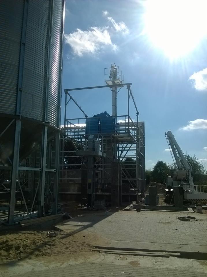 Budowa hali stalowej z silosami zbożowymi w słoneczny dzień. Widoczny żuraw budowlany i stalowa konstrukcja w trakcie montażu.