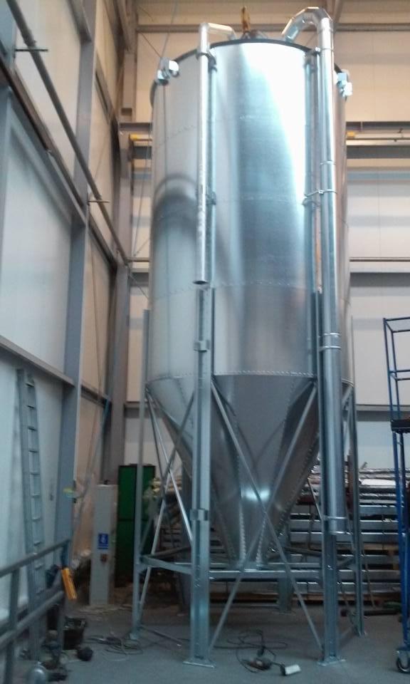 Wnętrze hali z metalową konstrukcją zbiornika na wysokich nogach, z widocznymi elementami mocującymi i rurami. W tle ściany hali i drabina.