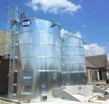 Dwa srebrne silosy zbożowe marki CIN, z drabinkami, na tle błękitnego nieba i fragmentu budynku. Widoczne detale konstrukcyjne i obudowa sterownicza.